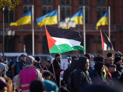 A person holds a Palestinian flag while standing in a public square flying Ukrainian flags during an outdoor rally