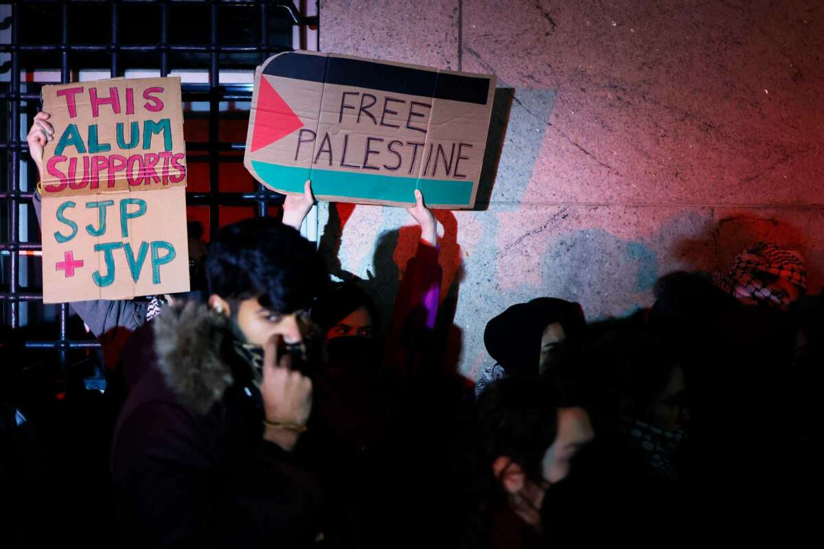 Two protesters hold signs - one reading "THIS ALUM SUPPORTS SJP + JVP" and the other reading "FREE PALESTINE" - while participating in an outdoor protest