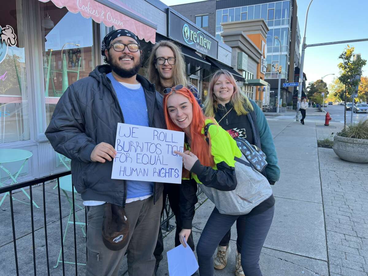 People stand outside of a restaurant to deliver an addressPeople pose for a photo around a sign that reads "WE ROLL BURRITOS TIGHT FOR EQUAL HUMAN RIGHTS" while standing on a city sidewalk