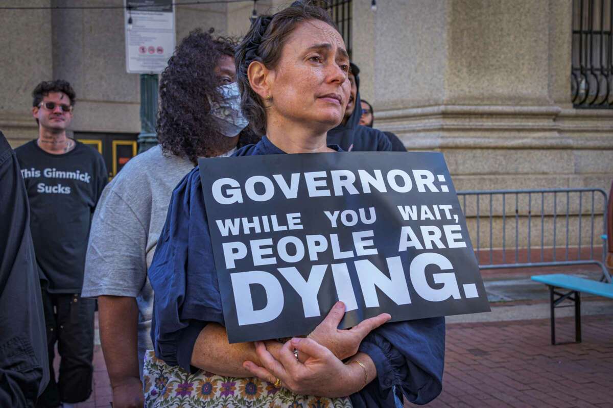 A protester holds a sign reading "GOVERNOR: WHILE YOU WAIT PEOPLE DIE" during an outdoor demonstration