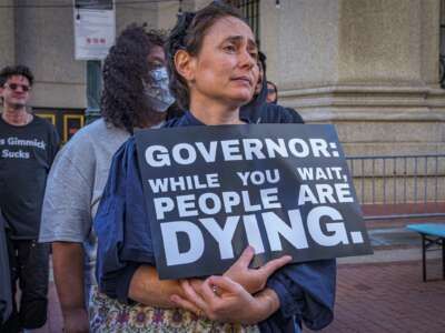 A protester holds a sign reading "GOVERNOR: WHILE YOU WAIT PEOPLE DIE" during an outdoor demonstration
