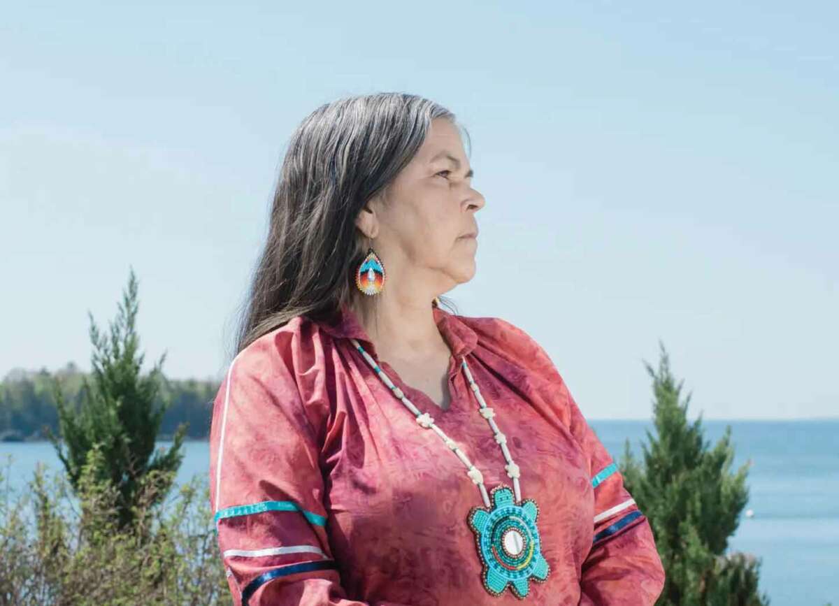 A woman wearing a light red tunic and an ornate beaded pendant stands outside among plants, and beside still water