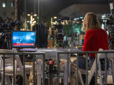 A woman in a red dress sits at a table opposite of several news cameras