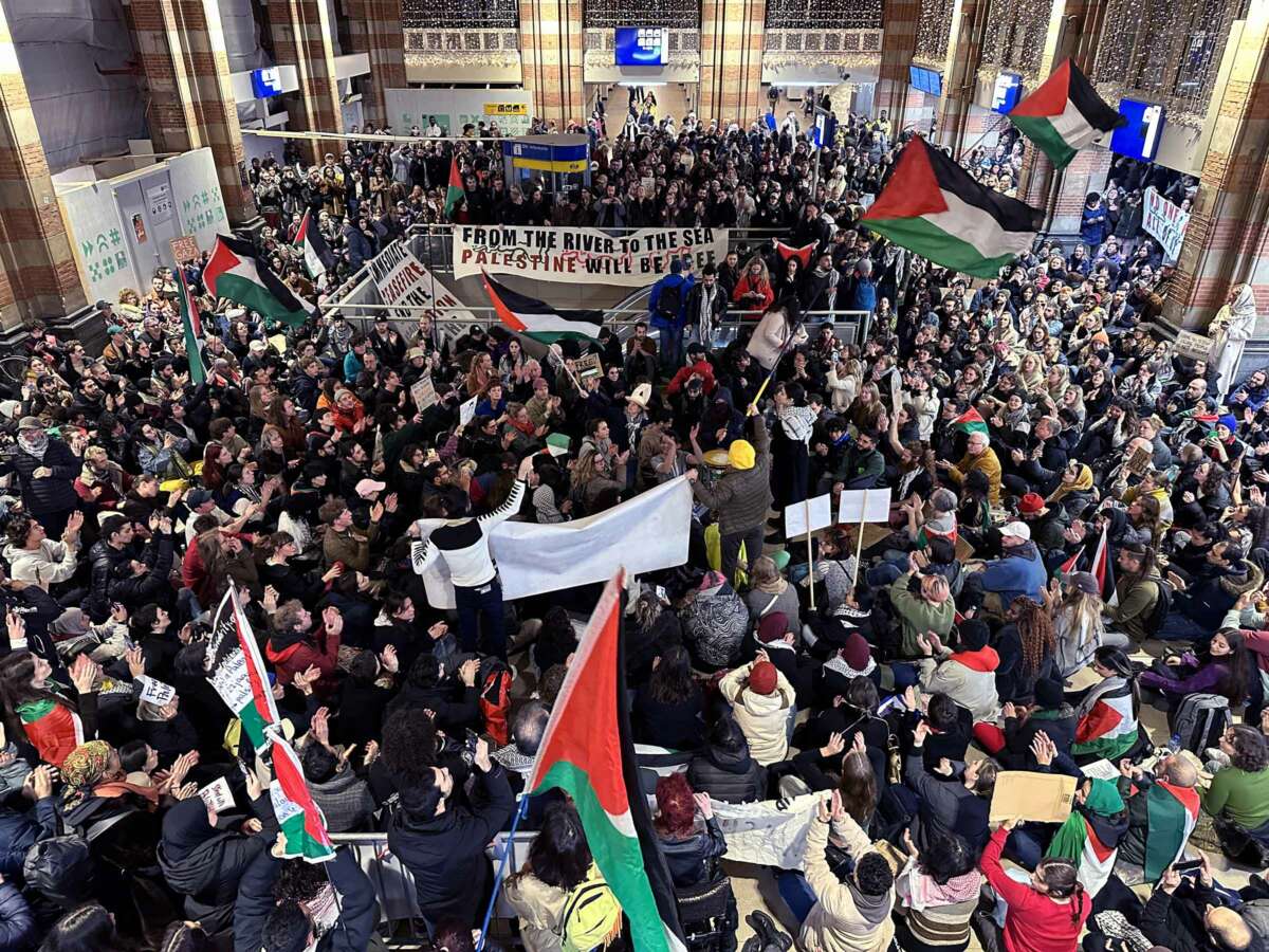 Hundreds of people waving the Palestinian flag and a banner reading "FROM THE RIVER TO THE SEA, PALESTINE WILL BE FREE" gather in an indoor space for a protest