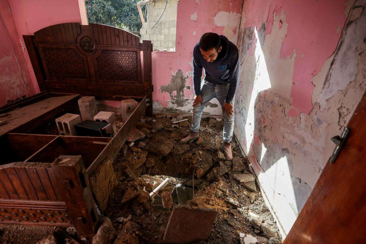 A man looks through a hole blown through his floor as he stands in a destroyed bedroom