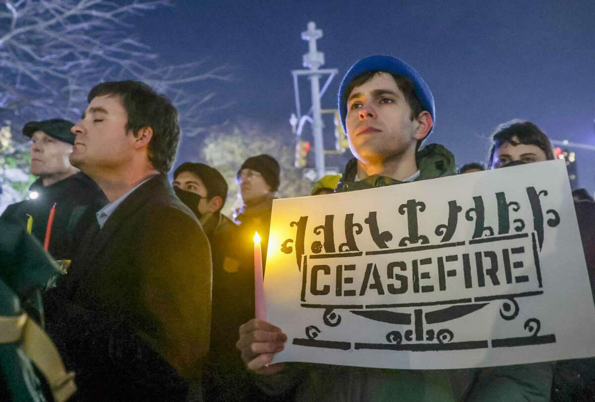 A man holds a sign with an image of a menorah with the word "CEASEFIRE" written across it during an outdoor demonstration