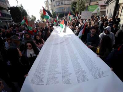 People gathered in a street for a demonstration carry a long list of the names of people martyred during Israeli attacks on Palestine