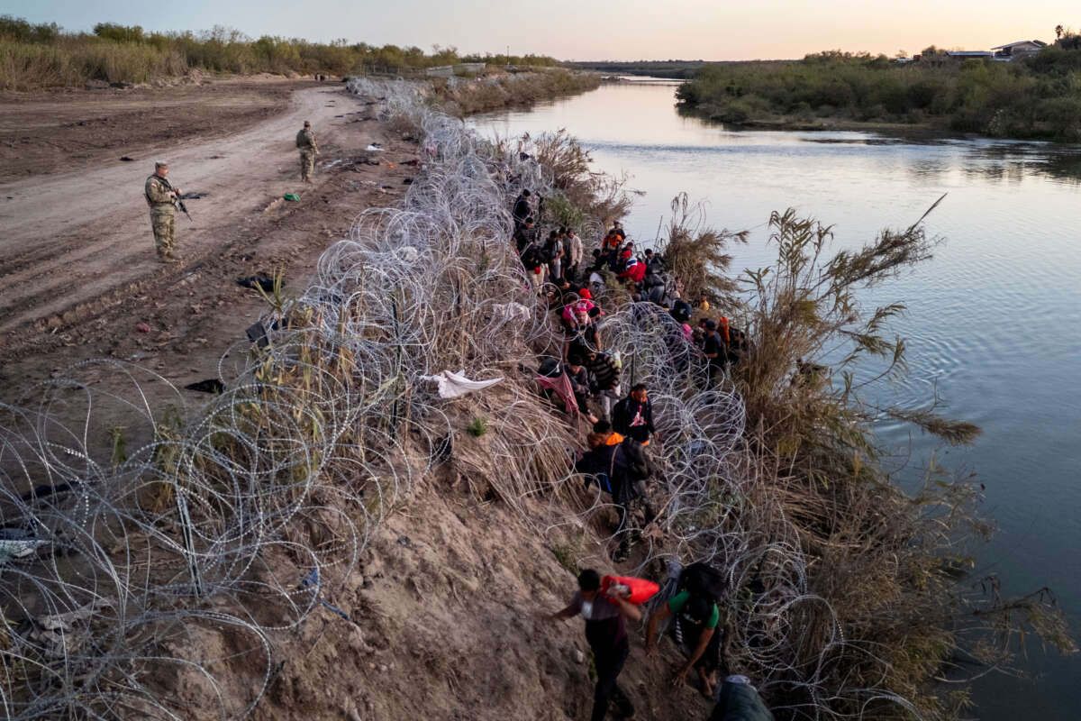 Texas National guardsmen watch as migrants pick their way through razor wire after crossing the Rio Grande into the United States on December 17, 2023, in Eagle Pass, Texas.