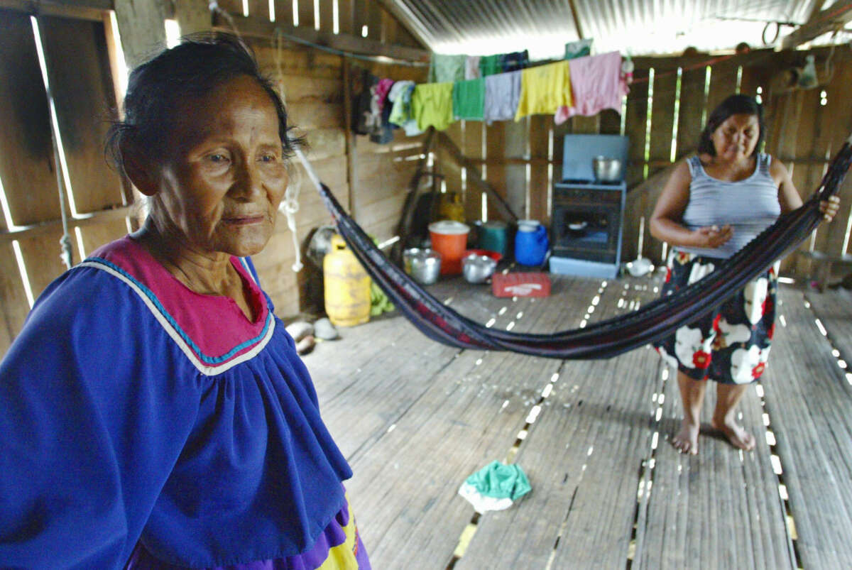 Two Indigenous women, one wearing traditional garments, stand in their home