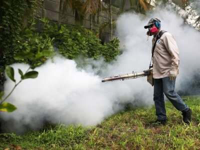 A mosquito control inspector wearing protective gear sprays a cloud of pesticide