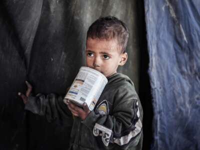 A little boy with soot on his face chews on the top of an unopened formula container