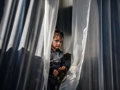 A little girl sits in a makeshift shelter made of a clear plastic curtain.