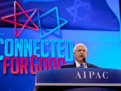 David Friedman speaks at a podium clearly labeled AIPAC with the words "CONNECTED FOR GOOD" alongside a 5-pointed star tangled with a Star of David behind him