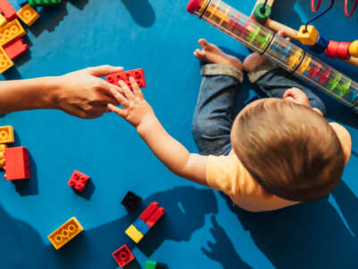 Person hands a child a block as child plays with toys in child care setting.