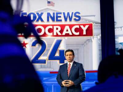 Ron DeSantis stands underneath a sign reading "FOX NEWS DEMOCRACY 2024" as he awaits being interviewed