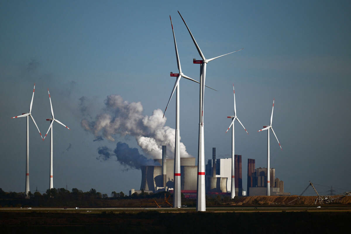 Wind turbines stand in the foreground against smokestacks billowing pollution into the air behind them