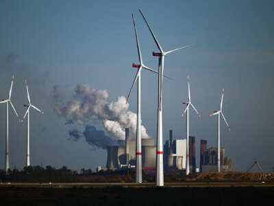 Wind turbines stand in the foreground against smokestacks billowing pollution into the air behind them