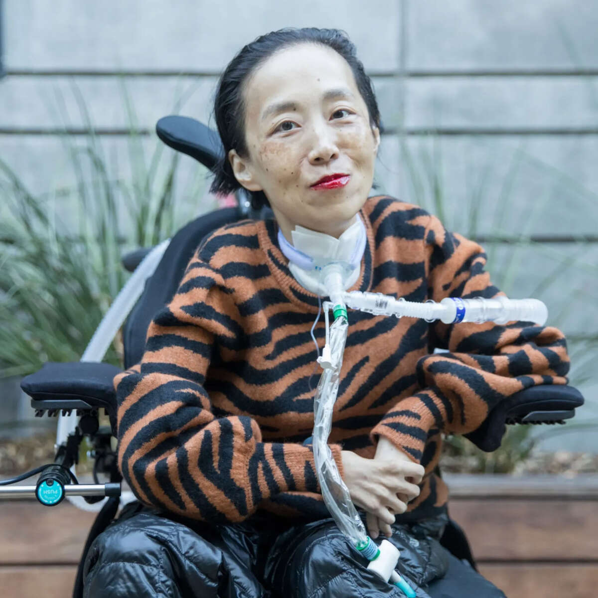 Image description: Photo of Alice Wong, an Asian American disabled woman in a power chair. She is wearing an orange and black tiger-striped sweater, black pants, a bold red lip color, and a trach at her neck. In the background is a gray cement wall with greenery.