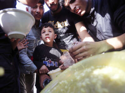 A boy stands among men, all of whom wait for soup with makeshift bowls in their hands.