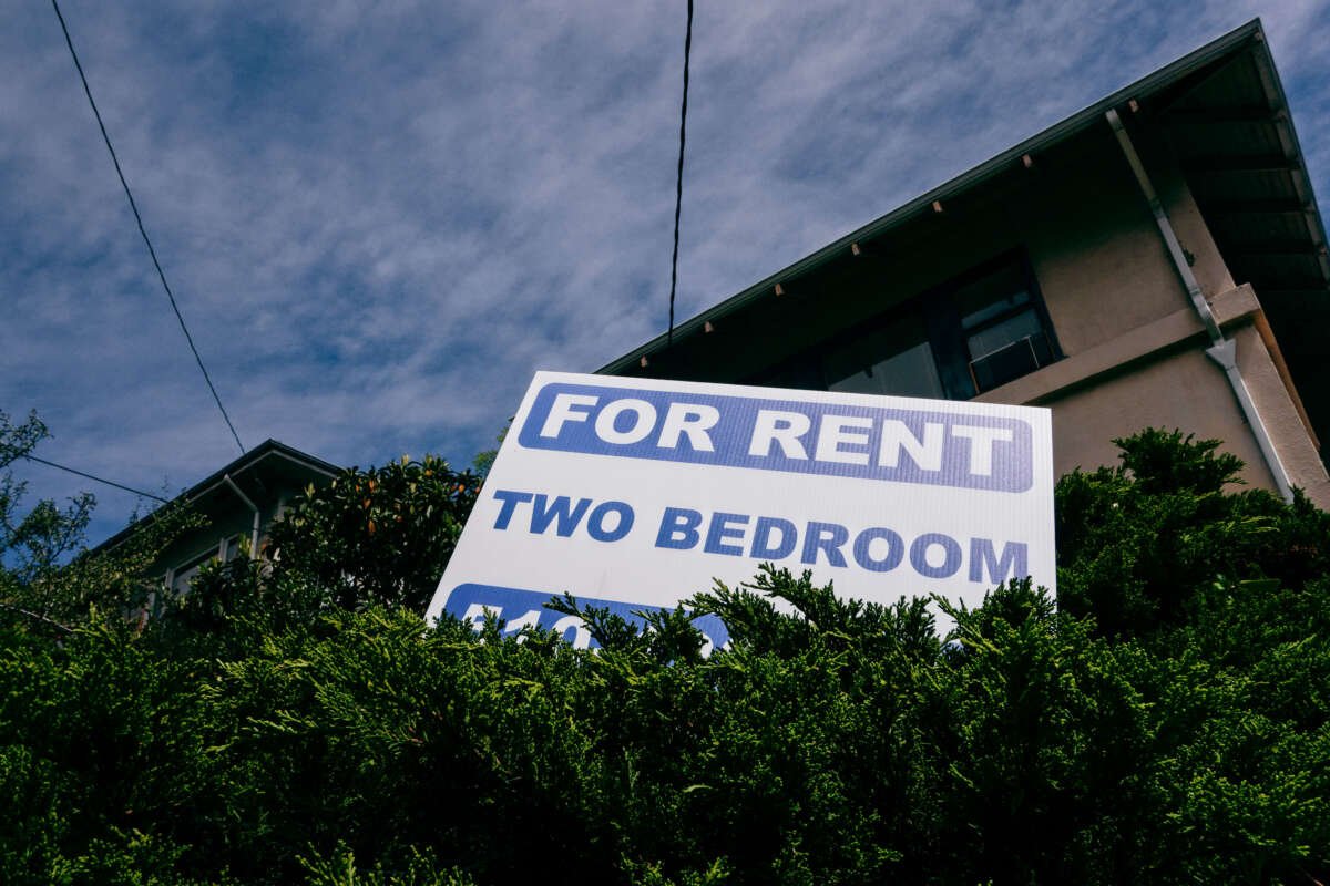 A sign reading 'For Rent / Two Bedroom" is seen in bushes in front of an apartment building, low angle.