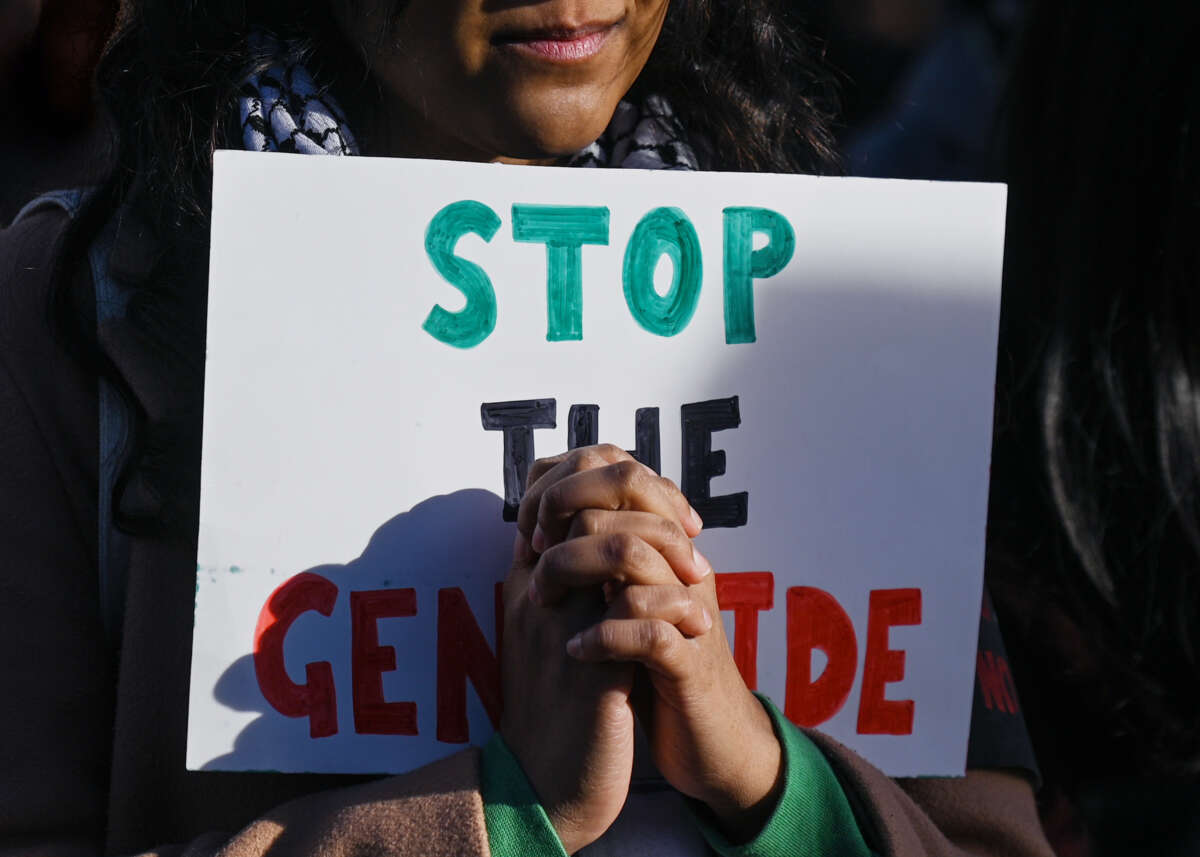 A person holds a sign reading "STOP THE GENOCIDE" behind their clasped hands
