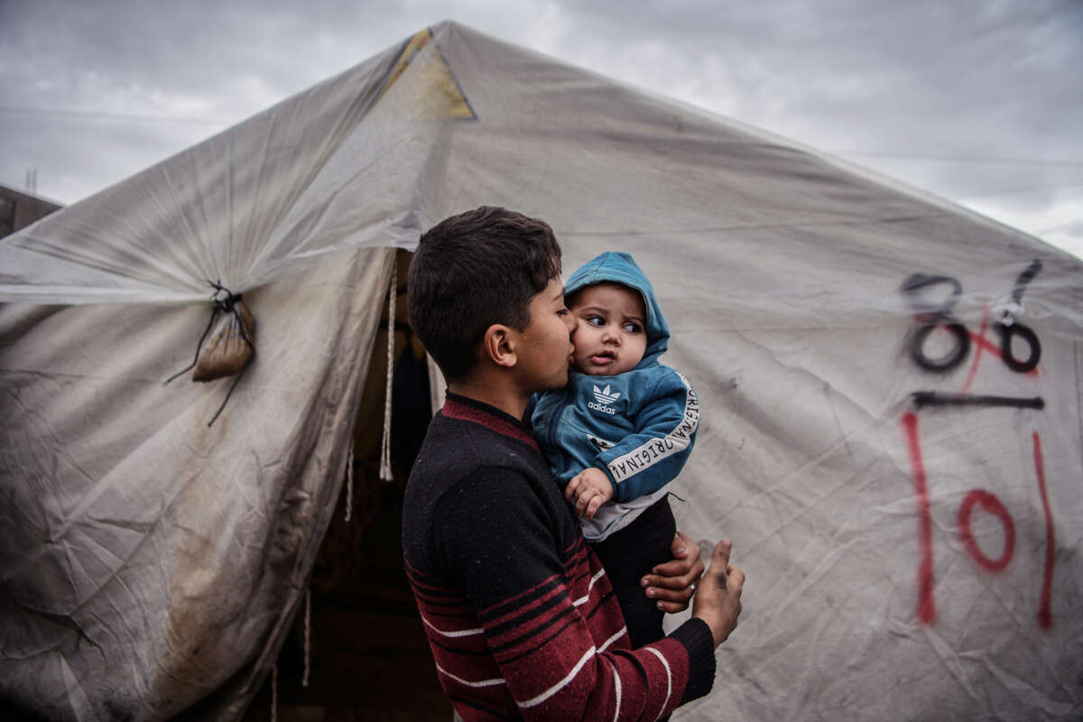 A teen boy kisses his infant brother on ths cheek as he holds him while standing outside of the tent they share in a refugee camp