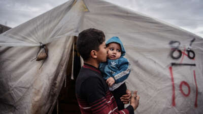 A teen boy kisses his infant brother on ths cheek as he holds him while standing outside of the tent they share in a refugee camp