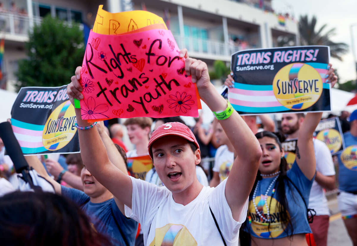 A protester holds a sign reading "RIGHTS FOR TRANS YOUTH" on a neon pink piece of paper as they march with others during an outdoor demonstration