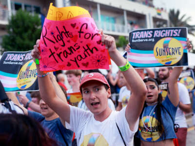 A protester holds a sign reading "RIGHTS FOR TRANS YOUTH" on a neon pink piece of paper as they march with others during an outdoor demonstration