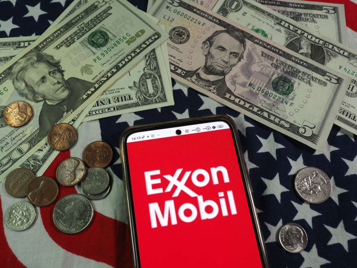 The Exxon Mobil Corporation logo is seen displayed on a smartphone with U.S. currency notes and coins in the background.