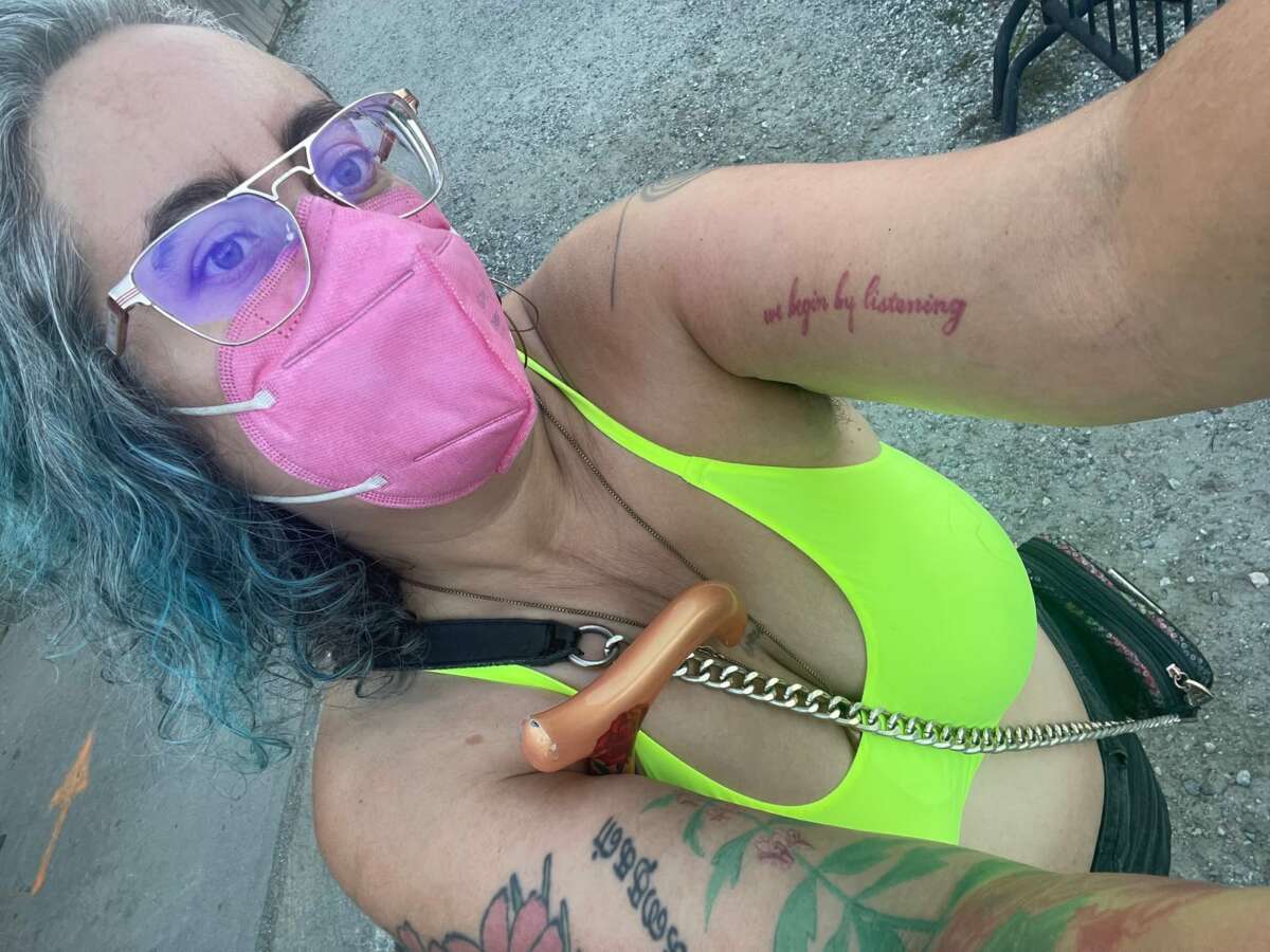 A photo of Leah Lakshmi Piepzna-Samarasinha, a non-binary femme of Sri Lankan and Irish/ Roma heritage with curly gray, brown, and blue hair. Leah is wearing a neon yellow halter top, a pink respirator mask, and metal-rimmed eyeglasses. They have a purse slung over their shoulder and a cane tucked under one arm as they hold their phone to take a selfie. They have visible tattoos on both arms. One of those tattoos reads, we begin by listening.