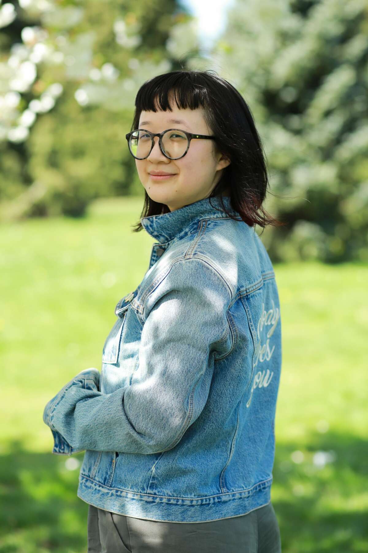 Photo of Jane Shi, a person with pale tan skin. Jane's body is facing away from the camera, but her face smiling towards it. She's wearing a blue jean jacket with the words "Nobody Loves You" embroidered in white across its back. Her hair is nearly shoulder-length with dark red highlights at the ends. She is wearing large, round, black-grey glasses. The background is a park with lawn grass and trees, out of focus.