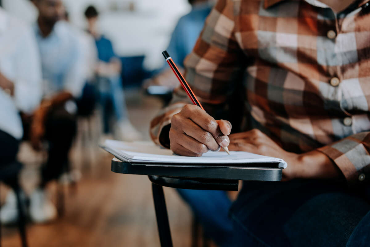 Black student writing in notebook on desk, close-up on hand with pencil