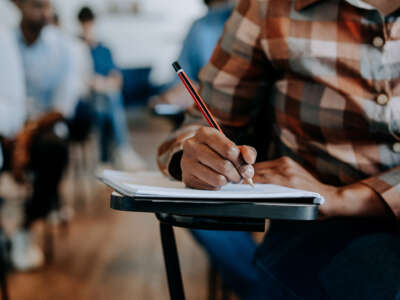 Black student writing in notebook on desk, close-up on hand with pencil