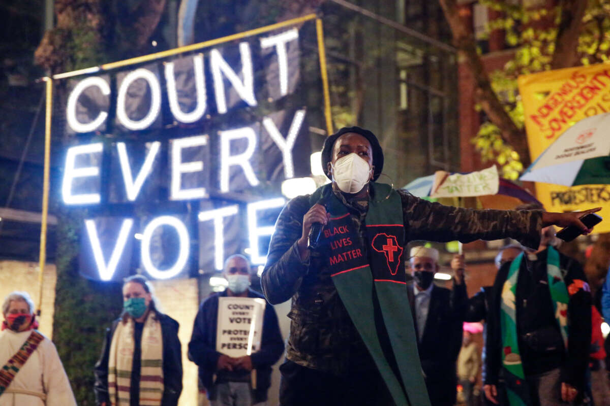 A protester in a ppe mask speaks into a microphone in front of a lit sign reading "COUNT EVERY VOTE"