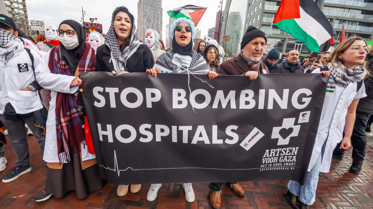 Medical professionals wearing Keffiyehs over their work uniforms march behind a banner reading: "STOP BOMBING HOSPITALS" during an outdoor protest