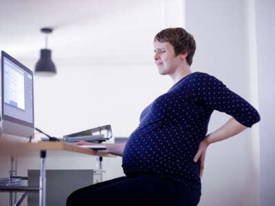 A pregnant person rubs their back while sitting at an office desk