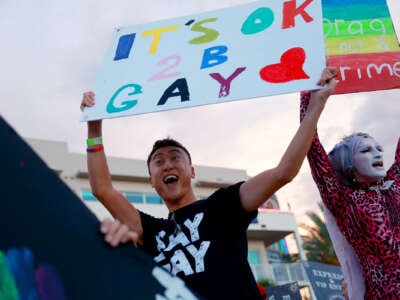 A jubliant person holds a sign reading "IT'S OK 2 B GAY" while wearing a shirt that reads "SAY GAY"