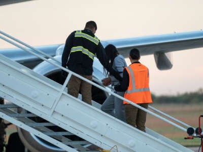 A person whose face is obscured by hair is led, handcuffed, into a plane by people wearing bright-colored visibility vests