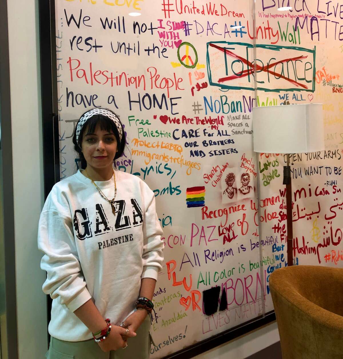 Intimaa Salama, in a gray sweatshirt that reads "GAZA", stands in front of a white board upon which many drawings and words are written.