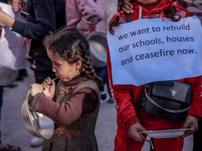 A child holds an empty bowl as another holds a sign reading "We want to rebuild our schools, houses; ceasefire now"