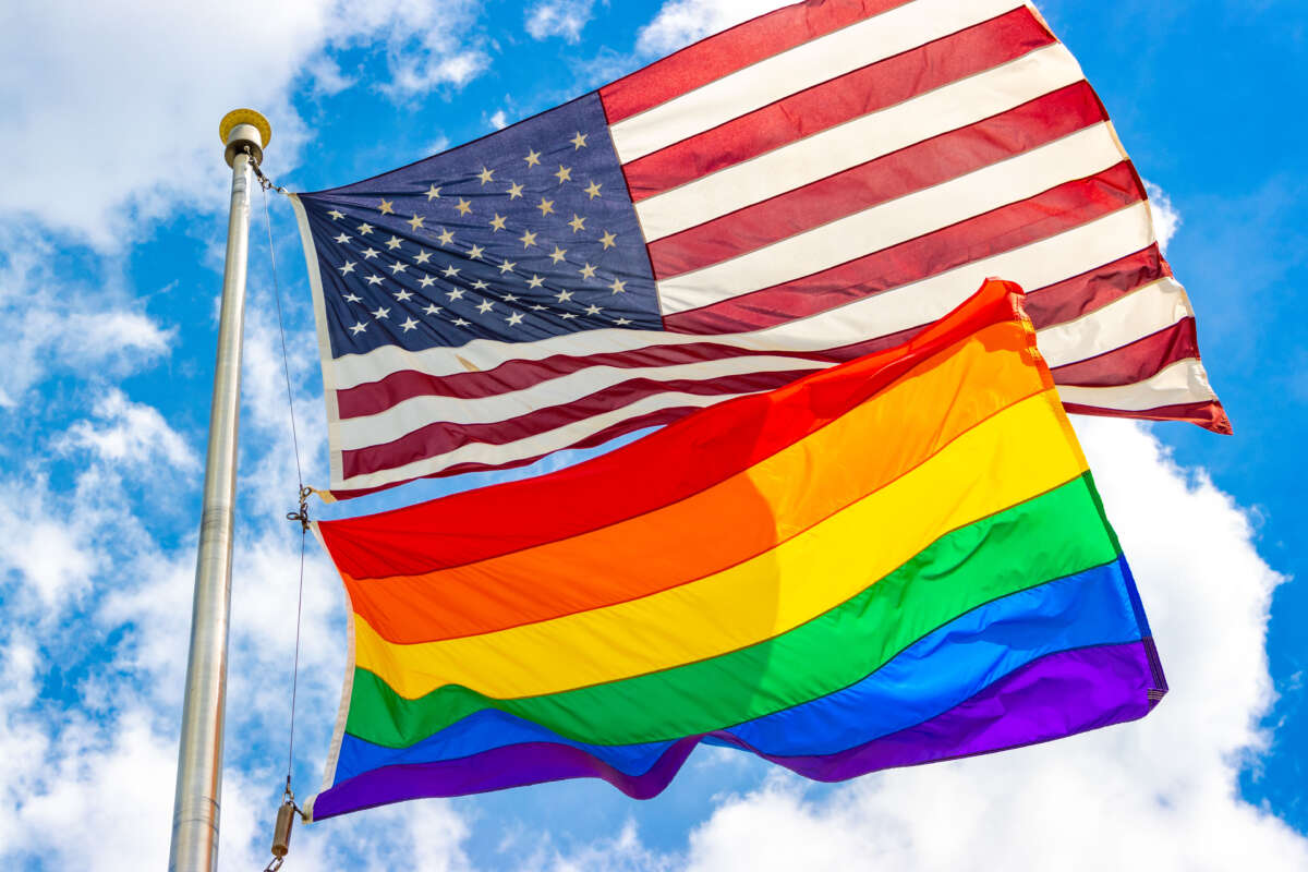 A USA flag and a pride flag fly over blue cloudy sky