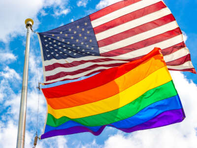 A USA flag and a pride flag fly over blue cloudy sky