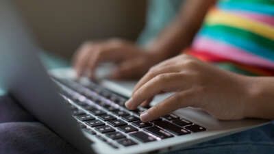 Youth with rainbow striped shirt types on laptop, close-up on hands on keys