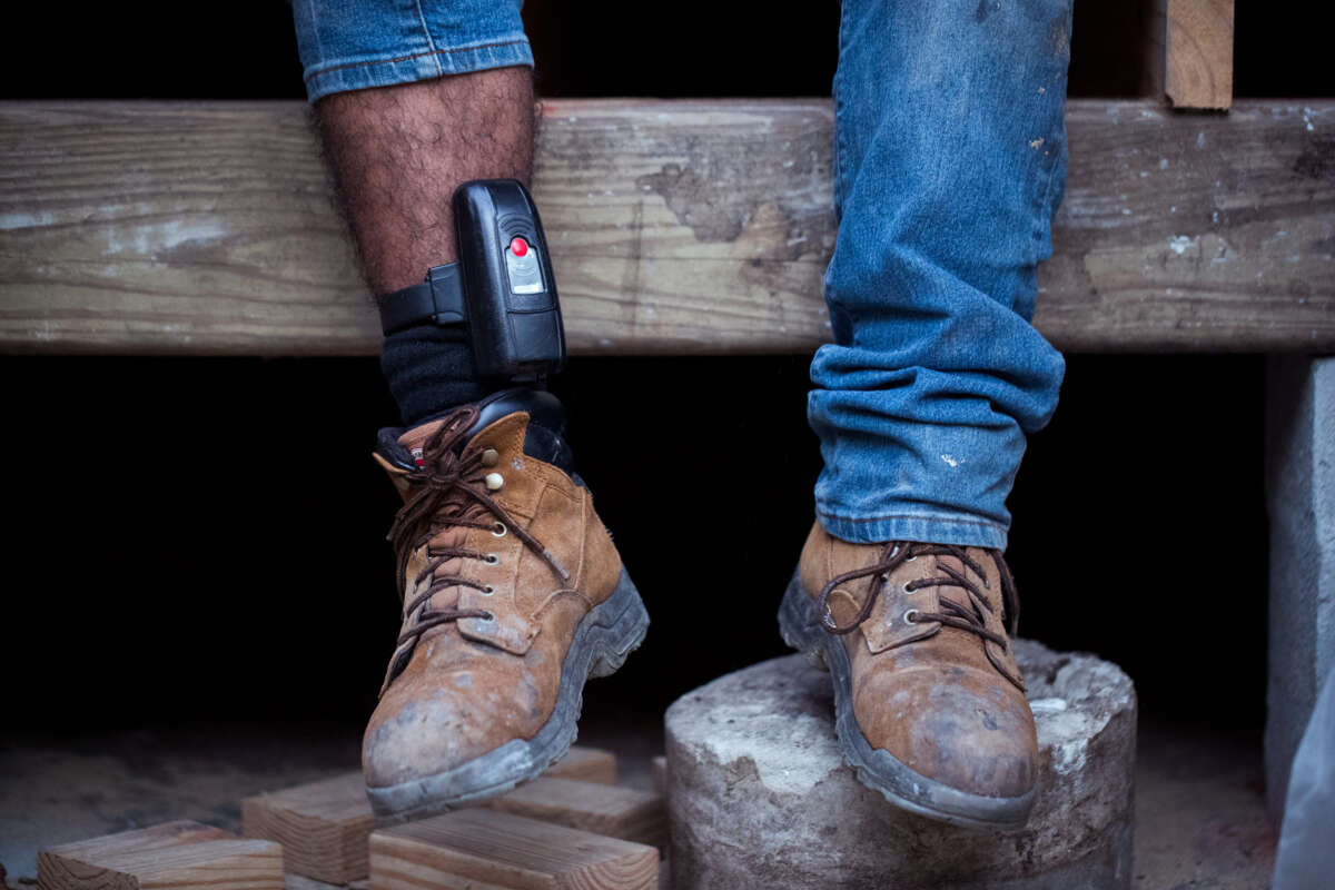 A man in work boots sits at a construction site with an ankle monitor locked around his right leg