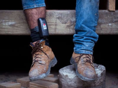 A man in work boots sits at a construction site with an ankle monitor locked around his right leg