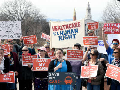 A woman in a blue dress holds a microphone while standing behind a podium reading "SAVE MY CARE" as others around her also hold signs