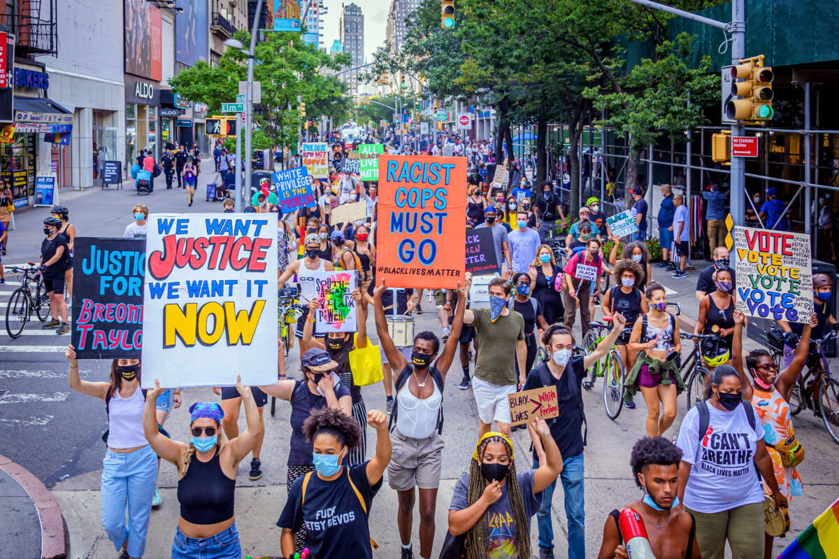 People march through the streets with colorful signs reading "WE WANT JUSTICE WE WANT IT NOW" and "RACIST COPS MUST GO" during a street protest