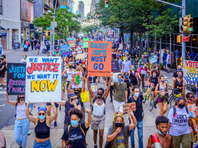 People march through the streets with colorful signs reading "WE WANT JUSTICE WE WANT IT NOW" and "RACIST COPS MUST GO" during a street protest