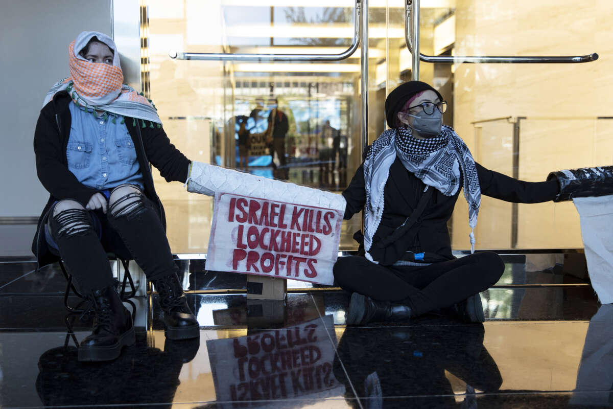 Activists with covered faces and lockboxed arms sit behind a sign reading "ISRAEL KILLS, LOCKHEED PROFITS" during a sit-in outside of the Lockheed Martin headquarters